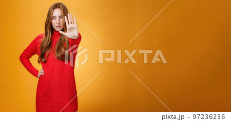 Confident sensual and attractive redhead female with self-assured gaze extending palm towards camera in stop and prohibition gesture demanding halt, stand still over orange background Confident sensual and attractive redhead female with self-assured gaze extending palm towards camera in stop and prohibition gesture demanding halt, stand still over orange background 97236236