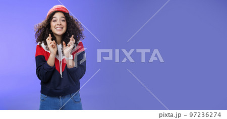 Optimistic and hopeful girl having faith in desire come true, standing with crossed fingers for good luck having wish anticipating miracle, smiling broadly at camera as wearing warm stylish red beanie 97236274