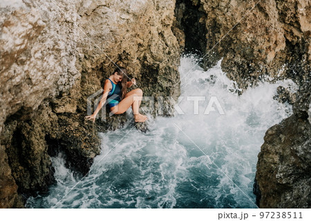 A young beautiful tanned woman with long hair in blue bikini sitting at rocks near blue green clear ocean sea water. Woman relax on sunset at sea, enjoy holidays and weekend vacation in summer time. A young beautiful tanned woman with long hair in blue bikini sitting at rocks near blue green clear ocean sea water. Woman relax on sunset at sea, enjoy holidays and weekend vacation in summer time. 97238511