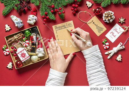 female hand holds pen and writing Christmas shopping list, gift ideas on white notepad on red background Top view Flat lay Holiday card Woman making Christmas presents list, Planning, buying concept. 97239124