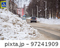 A large, high snowdrift against the backdrop of a city street with cars.  97241909