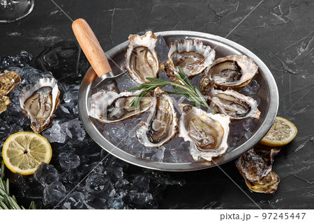 Fresh opened oysters, ice and lemon on a round metal plate, black stone textured background. Top view. Close-up shot. 97245447