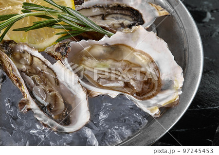 Fresh opened oysters, ice and lemon on a round metal plate, black stone textured background. Top view. Close-up shot. Fresh opened oysters, ice and lemon on a round metal plate, black stone textured background. Top view. Close-up shot. 97245453