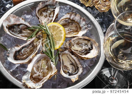 Fresh opened oysters, ice, lemon on a round metal plate and champagne are on a black stone textured background. Top view. Close-up shot. 97245454