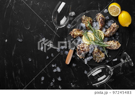 Fresh closed oysters, ice, lemon on a round slate and two empty glasses are on a black stone textured background. Top view with copy space. Close-up. Fresh closed oysters, ice, lemon on a round slate and two empty glasses are on a black stone textured background. Top view with copy space. Close-up. 97245483