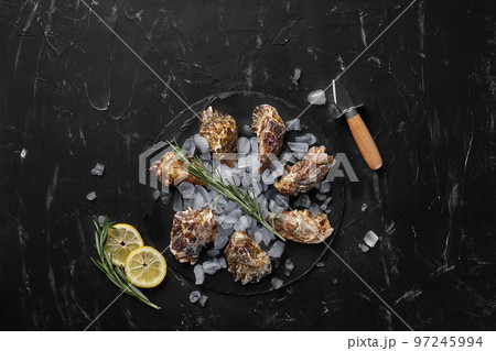 Fresh closed oysters, ice and lemon on a round slate, black stone textured background. Top view with copy space. Close-up shot. Fresh closed oysters, ice and lemon on a round slate, black stone textured background. Top view with copy space. Close-up shot. 97245994