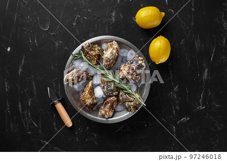 Fresh closed oysters, ice and lemon on a round metal plate, black stone textured background. Top view with copy space. Close-up shot. Fresh closed oysters, ice and lemon on a round metal plate, black stone textured background. Top view with copy space. Close-up shot. 97246018