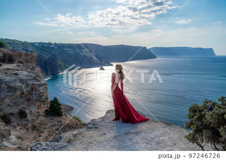 A girl with flowing hair in a long red dress stands on a rock above the sea. The stone can be seen in the sea. Sunny path to the sea from the sun. A girl with flowing hair in a long red dress stands on a rock above the sea. The stone can be seen in the sea. Sunny path to the sea from the sun. 97246726