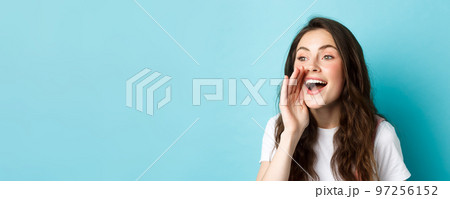 Close up portrait of cheerful young woman calling someone, holding hand near opened mouth while shouting, searching for person, speaking loud, standing over blue background 97256152