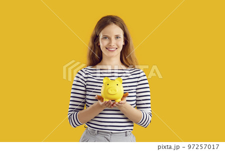 Portrait of positive woman in striped sweatshirt holding piggy bank in hands on yellow background. 97257017