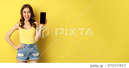 Stylish smiling woman showing mobile phone screen, app interface on smartphone, standing in tank top and shorts against yellow background 97257457