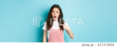 Surprised girl gasping and pointing at herself, look with disbelief, being picked or named, staring amazed at camera, standing against blue background 97257609