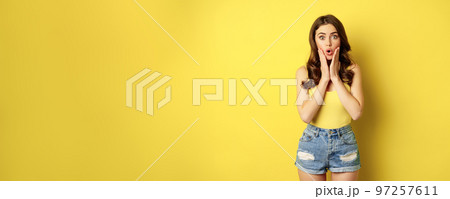 Portrait of brunette woman reacting surprised to advertsement, gasping and saying wow, wearing summer clothes, standing over yellow background 97257611