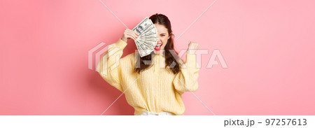 Excited beautiful girl saying yes, earn prize money, holding dollar bills on half of face, making fist pump gesture with satisfied expression, standing against pink background 97257613