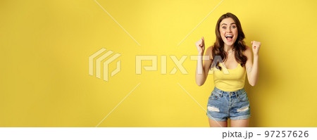 Enthusiastic young woman shouting and cheering, rejoicing, raising hands up and screaming in joy, chanting, standing over yellow background Enthusiastic young woman shouting and cheering, rejoicing, raising hands up and screaming in joy, chanting, standing over yellow background 97257626