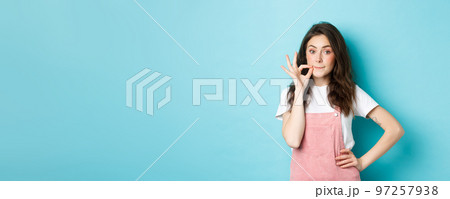 Cute glamour girl making lips zipper gesture, lock her mouth, promise keep secret, standing against blue background Cute glamour girl making lips zipper gesture, lock her mouth, promise keep secret, standing against blue background 97257938
