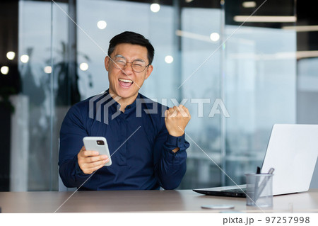 Portrait of happy Asian businessman, man celebrating achievement good results and victory, boss looking at camera inside office holding phone, entrepreneur working inside office. Portrait of happy Asian businessman, man celebrating achievement good results and victory, boss looking at camera inside office holding phone, entrepreneur working inside office. 97257998