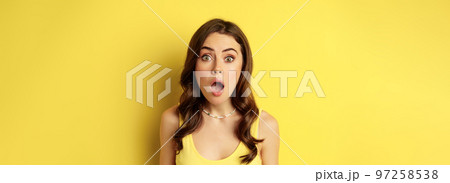 Close up portrait of stylish brunette girl looking amazed, wow face, showing excitement and surprise, impressed by something, standing over yellow background Close up portrait of stylish brunette girl looking amazed, wow face, showing excitement and surprise, impressed by something, standing over yellow background 97258538