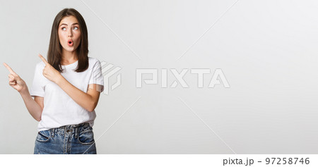 Excited attractive brunette girl in white t-shirt pointing fingers upper left corner, looking curious 97258746