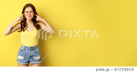 Young stylish woman feeling discomfort from loud noise sound, shut ears with fingers and complaining, standing over yellow background Young stylish woman feeling discomfort from loud noise sound, shut ears with fingers and complaining, standing over yellow background 97258919