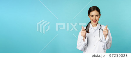 Enthusiastic woman doctor smiling, pointing fingers at camera, congratulating, inviting patients to clinic, standing over torquoise background Enthusiastic woman doctor smiling, pointing fingers at camera, congratulating, inviting patients to clinic, standing over torquoise background 97259023