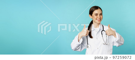 Happy woman doctor, medical worker in white coat, showing thumbs up in approval, like something, praise, standing over blue background Happy woman doctor, medical worker in white coat, showing thumbs up in approval, like something, praise, standing over blue background 97259072
