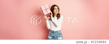 Cute smiling girl guessing whats inside pink present box, shaking gift and laughing, standing against studio background Cute smiling girl guessing whats inside pink present box, shaking gift and laughing, standing against studio background 97259094