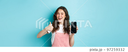Yes this is good. Smiling cute girl showing thumb up and empty smartphone screen, recommending application or online store, standing over blue background 97259332