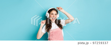 Image of cheerful young pretty girl dancing in headphones, listening music in app and jumping excited, standing over blue background 97259337