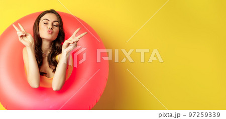 Portrait of attractive modern girl inside trendy pink beach swimming ring, showing peace v-sign and smiling happy, standing over yellow background Portrait of attractive modern girl inside trendy pink beach swimming ring, showing peace v-sign and smiling happy, standing over yellow background 97259339