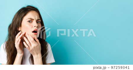 Skin care. Image of worried teenage girl looking in mirror and spot a pimple on face, trying squeeze acne on cheek, frowning upset, standing over blue background 97259341