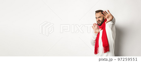 Guy asking to stop, grimacing and pulling away from camera, covering face with hands, standing over white background in red scarf and sweater 97259551