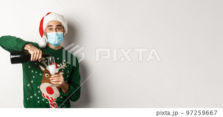 Concept of covid-19 and Christmas holidays. Happy guy in face mask and santa hat, celebrating New Year party and pouring glass of champagne, white background 97259667