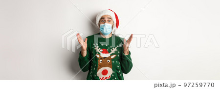 Christmas during pandemic, covid-19 concept. Surprised guy in medical mask, Santa hat and sweater celebrating New Year party, standing over white background Christmas during pandemic, covid-19 concept. Surprised guy in medical mask, Santa hat and sweater celebrating New Year party, standing over white background 97259770