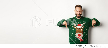 Winter holidays and christmas. Happy caucasian man in green sweater, pointing fingers down and looking satisfied, standing over white background 97260290
