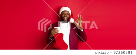 Amazed Black man holding holiday presents inside christmas sock, showing ok sign in approval, like something, standing over red background Amazed Black man holding holiday presents inside christmas sock, showing ok sign in approval, like something, standing over red background 97260291