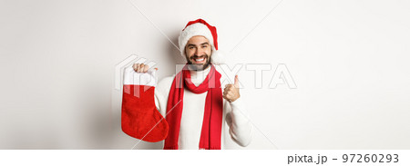 Winter holidays and celebration concept. Happy man showing Christmas sock for gifts and thumb up, smiling satisfied, standing over white background 97260293