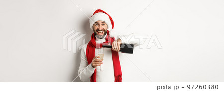 Christmas party and holidays concept. Happy bearded man in Santa hat and scard, pour himself champagne and smiling, celebrating New Year, standing over white background Christmas party and holidays concept. Happy bearded man in Santa hat and scard, pour himself champagne and smiling, celebrating New Year, standing over white background 97260380