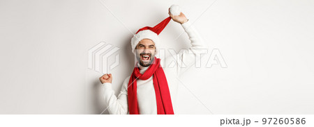 Winter holidays and New Year party concept. Happy man celebrating Christmas dancing in Santa hat and red scarf, standing against white background 97260586