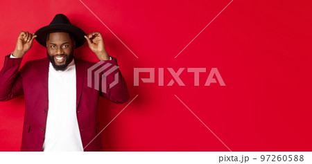 Fashion and party concept. Handsome african american man wearing hat and jacket, smiling self-assured, standing against red background 97260588