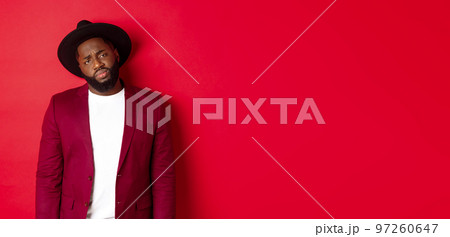 Sad and unamused Black man looking with pity at camera, frowning disappointed, standing against red background 97260647