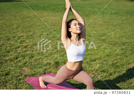 Portrait of asian young woman smiling pleased, stretching her body in yoga asana, standing on mat and workout in park Portrait of asian young woman smiling pleased, stretching her body in yoga asana, standing on mat and workout in park 97260831