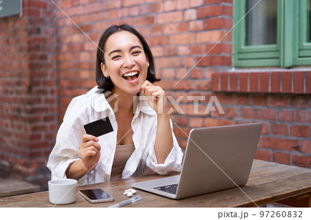 Happy young asian woman sitting near laptop, holding credit card, paying bills, shopping online contactless, smiling at camera Happy young asian woman sitting near laptop, holding credit card, paying bills, shopping online contactless, smiling at camera 97260832