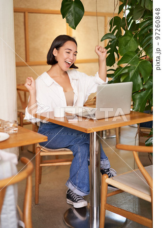 Enthusiastic asian girl celebrating, looking at laptop and triumphing, makes fist pumps with excited face, sits in cafe Enthusiastic asian girl celebrating, looking at laptop and triumphing, makes fist pumps with excited face, sits in cafe 97260848