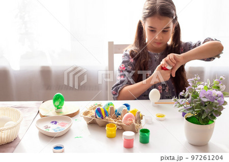 Colorful painted eggs in a tray stand on the table, the girl opens a jar of paint in the background. 97261204