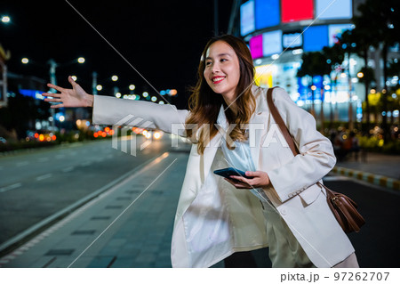 Beautiful woman smiling using smartphone application hailing with hand up calling cab after late work, Asian businesswoman standing hail waving hand taxi on road in busy city street at night 97262707