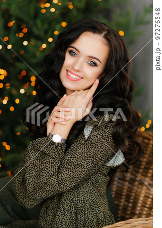 A smiling attractive brunette with professional makeup with her eyes closed against the background of lights holds her hands near her face 97276548