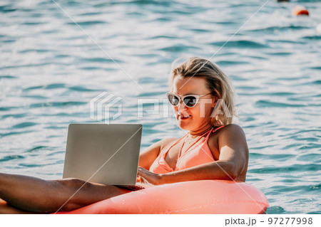 Woman works on laptop in sea. Freelancer, young blond woman in sunglases floating on an inflatable big pink donut with a laptop in the sea at sunset. Freelance, travel and holidays concept. 97277998
