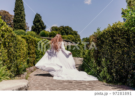 a beautiful woman with long brown hair and long white dress runs along a path along beautiful bushes in the park rear view 97281891