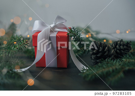 Red gift box with white bow on the background of fir branches and garland lights on a wooden table. Red gift box with white bow on the background of fir branches and garland lights on a wooden table. 97283105